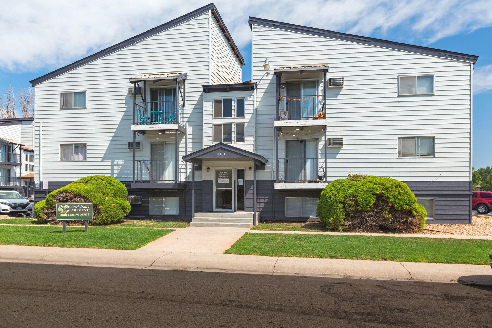 Apartment building exterior, white siding, black trim. Entryway, balconies, green bushes, and a blue sky.