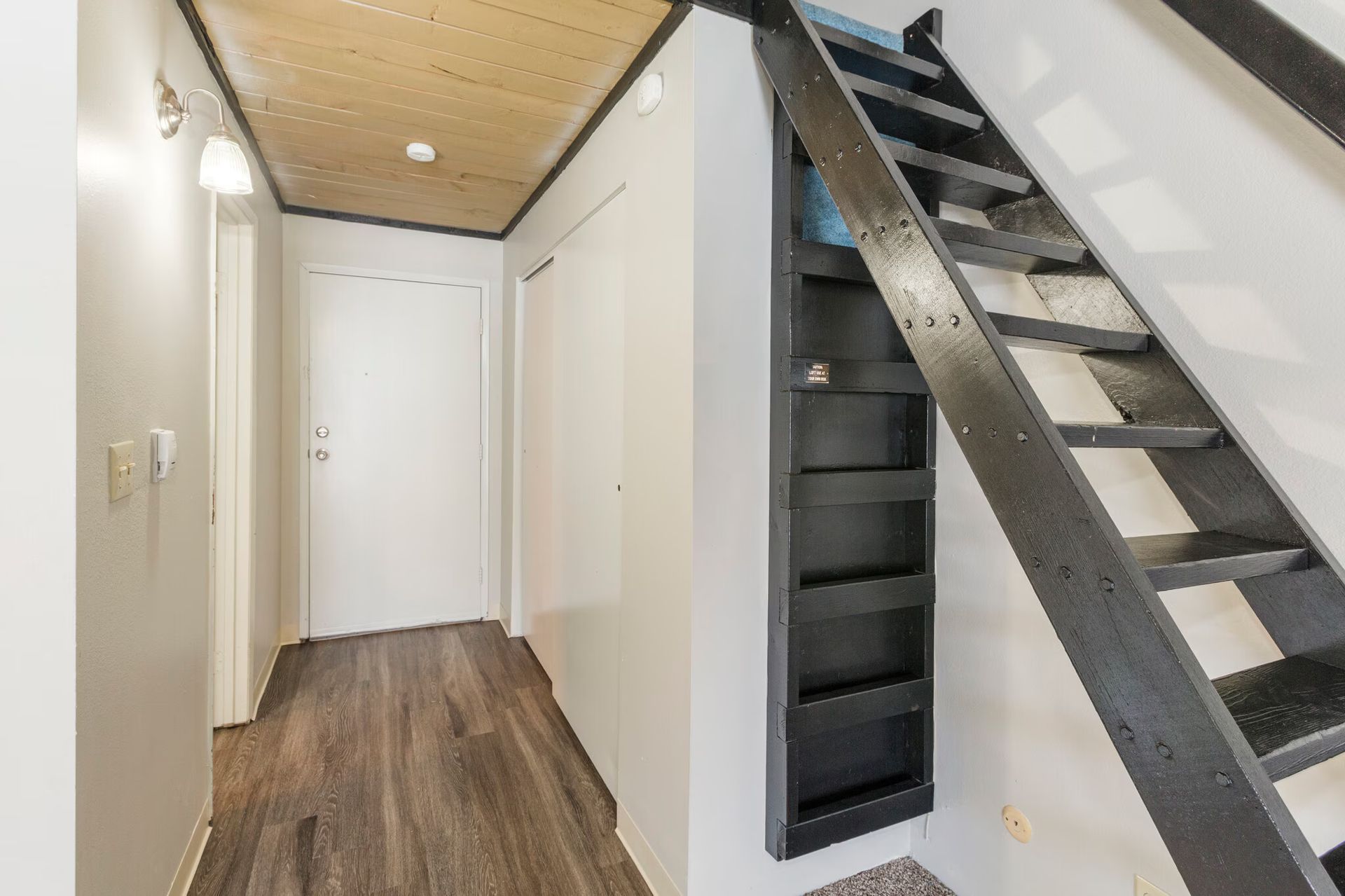 Hallway with a wooden floor, white walls, and a black ladder leading upwards.