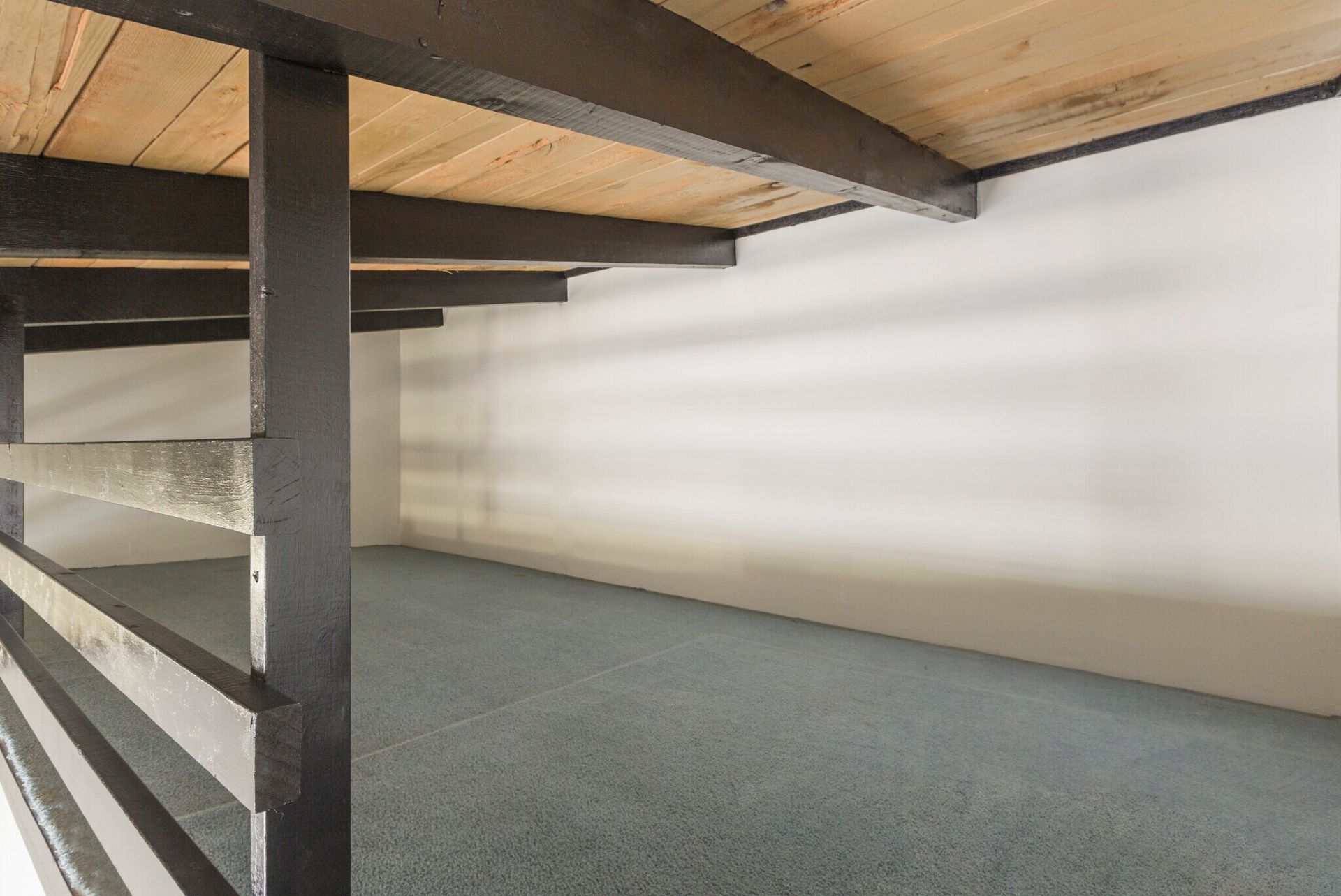 Empty space under a loft bed with dark supports and gray carpet, white wall.