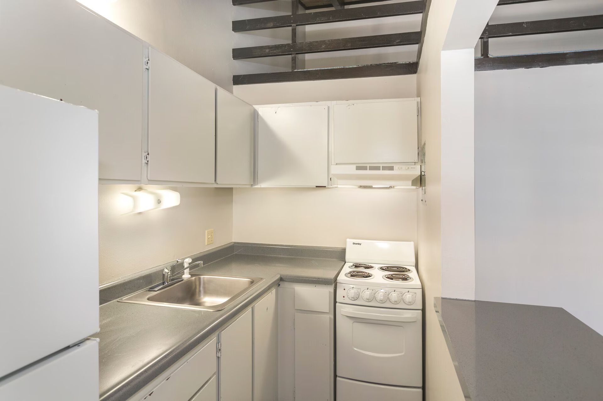 Small white kitchen with cabinets, stove, and sink. Dark countertops and a light fixture.