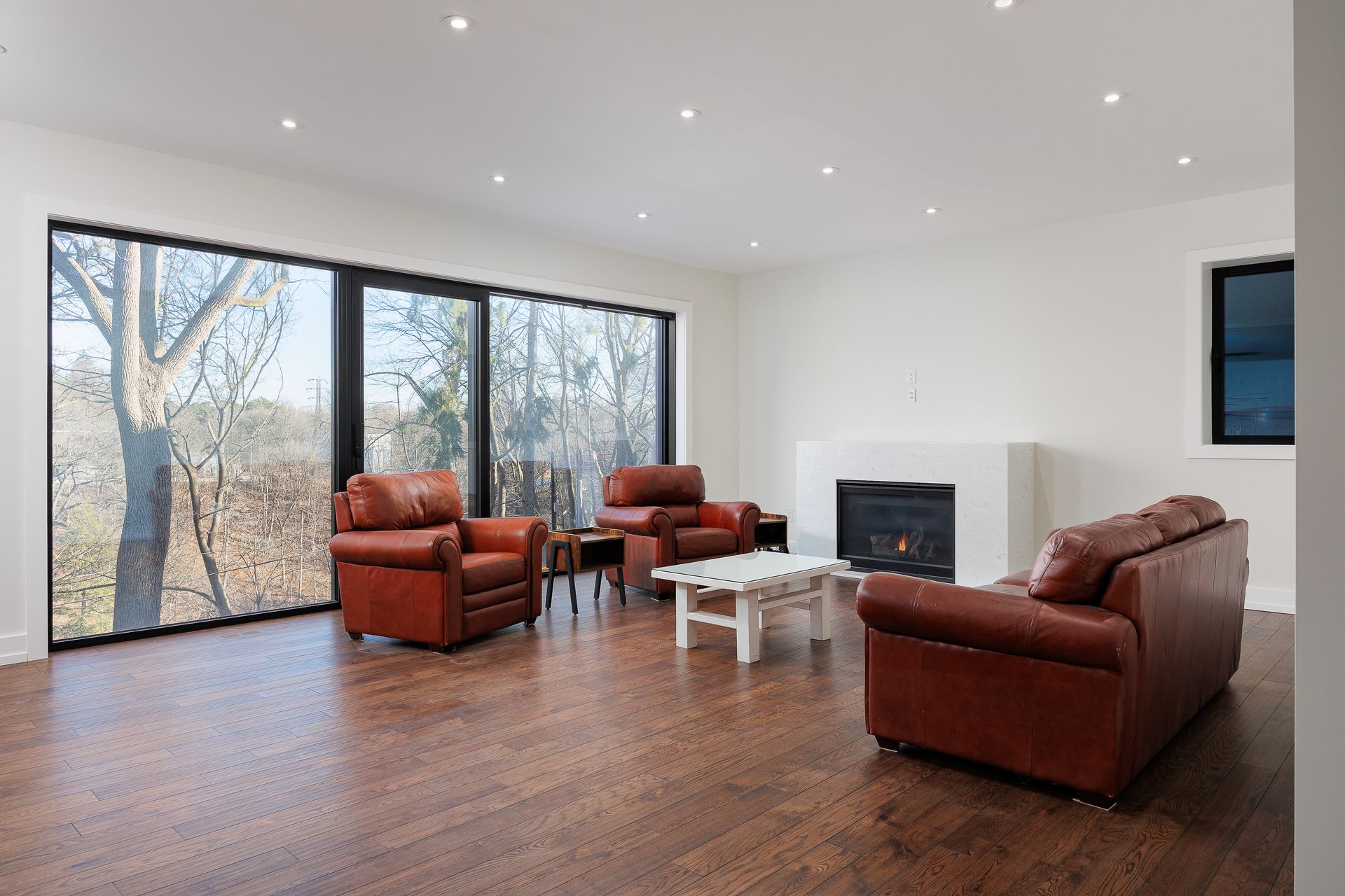 Modern living room with brown leather chairs, white tables, fireplace, and large windows.