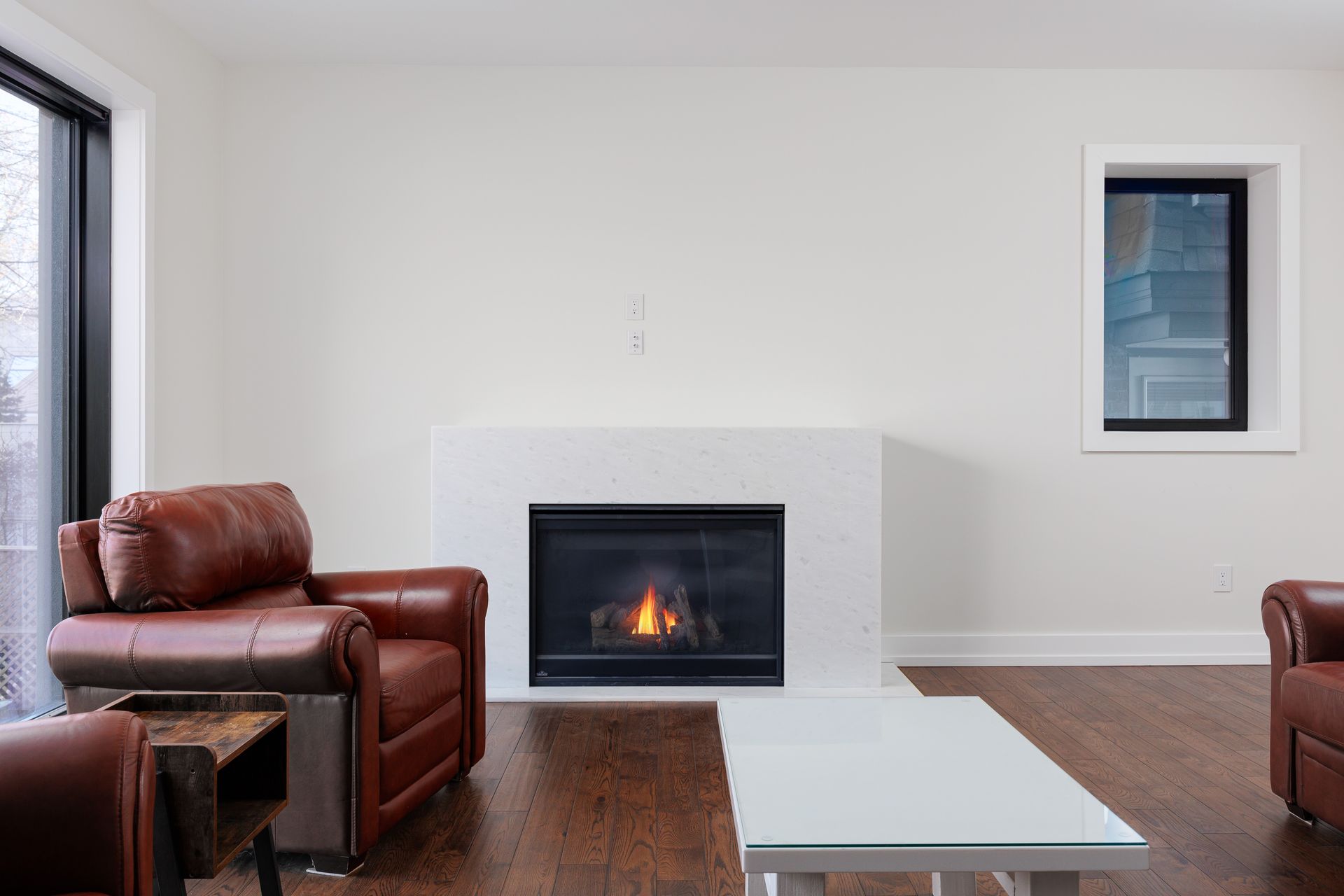 Modern living room with a lit fireplace, leather chairs, and a low white coffee table on dark wood floors