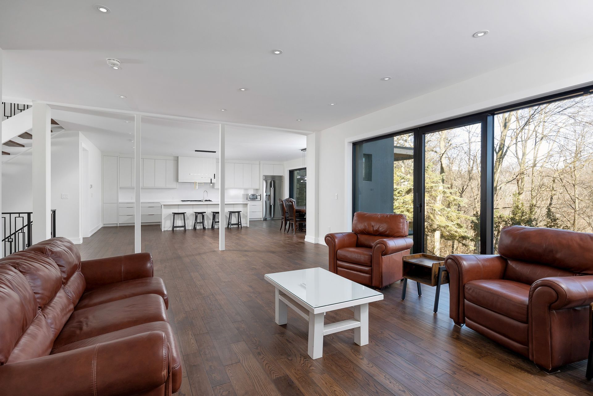 Modern living room with brown leather seating, white coffee table, and large windows overlooking trees