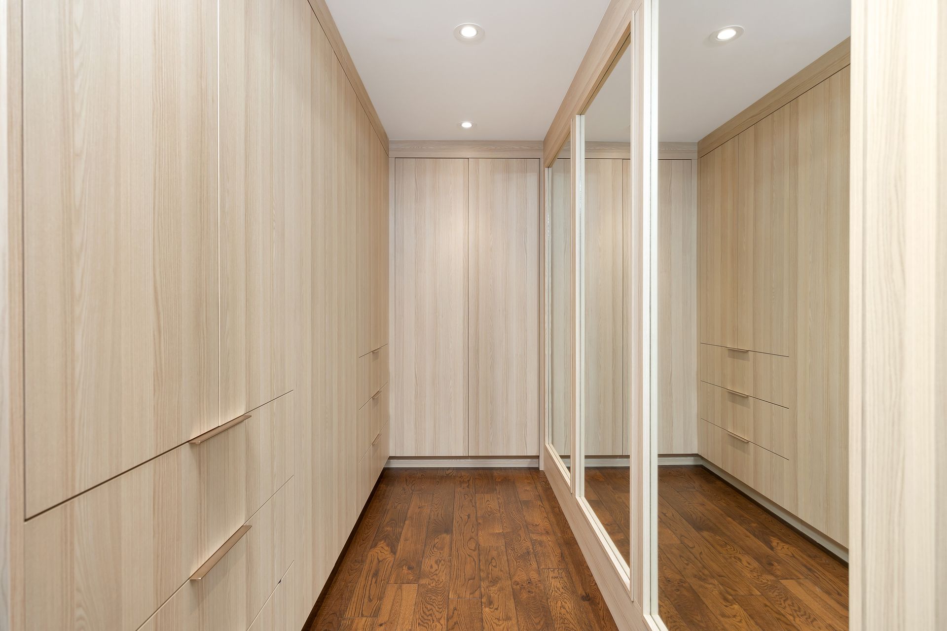 Narrow walk-in closet with light wood cabinetry, mirrored doors, and hardwood floor