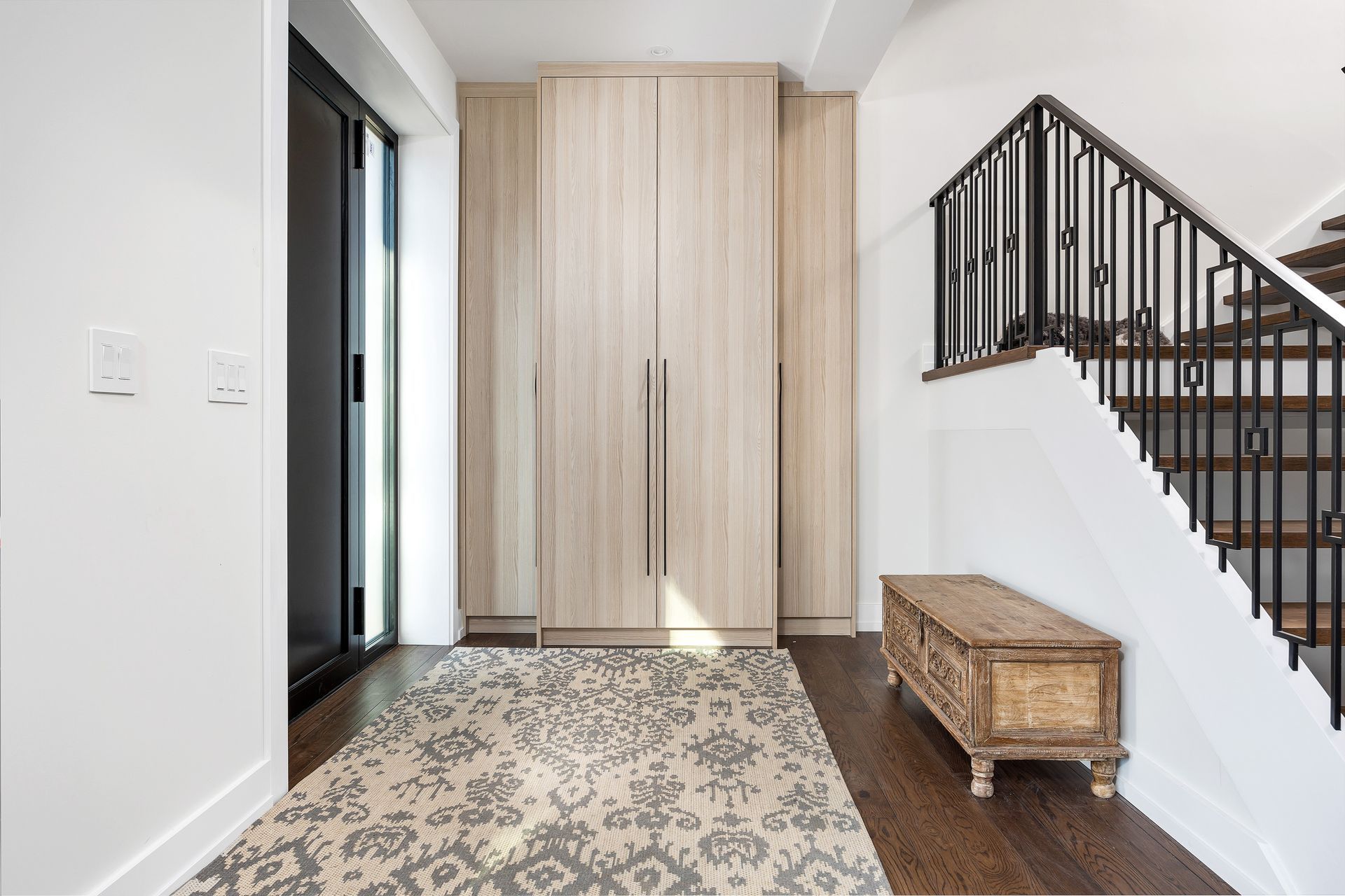Modern entryway with a wood wardrobe, patterned rug, dark floor, and staircase railing.