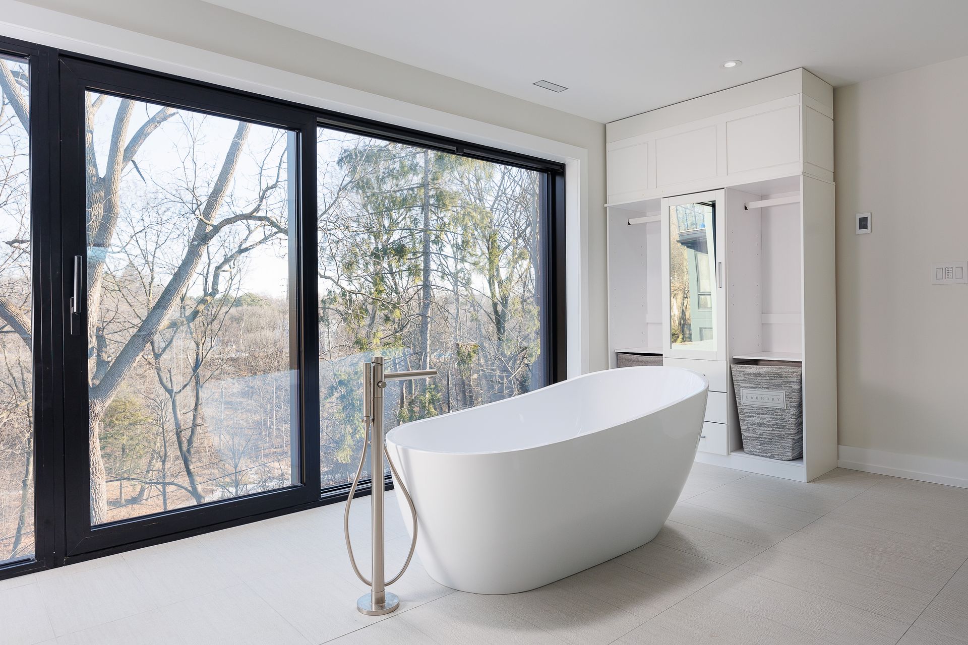 Modern bathroom with freestanding tub, large black-framed windows, and white cabinetry overlooking trees