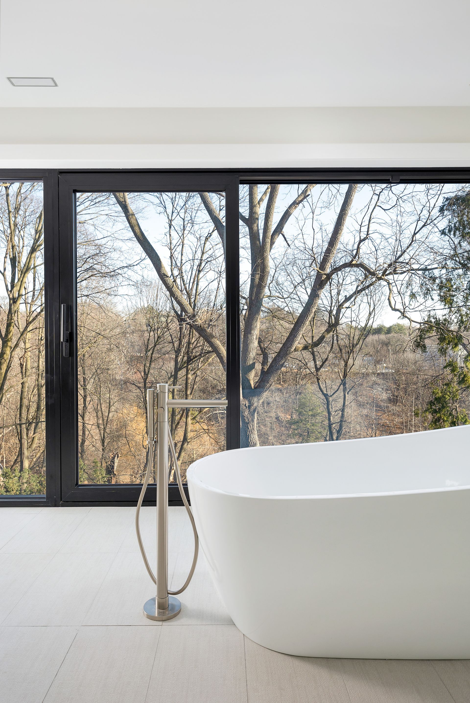 Modern bathroom with a freestanding white tub beside large black-framed windows and trees outside