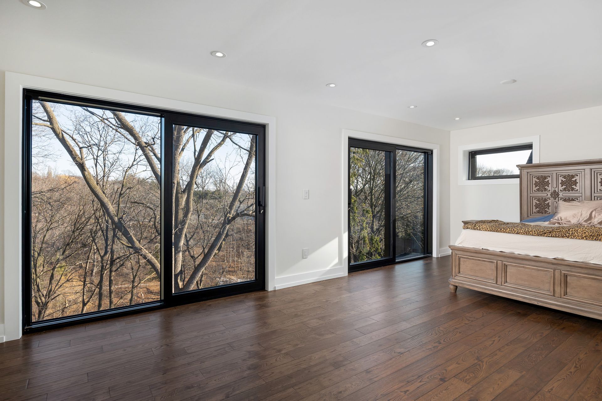 Modern bedroom with large windows, dark wood floors, white walls, and a bed with patterned bedding