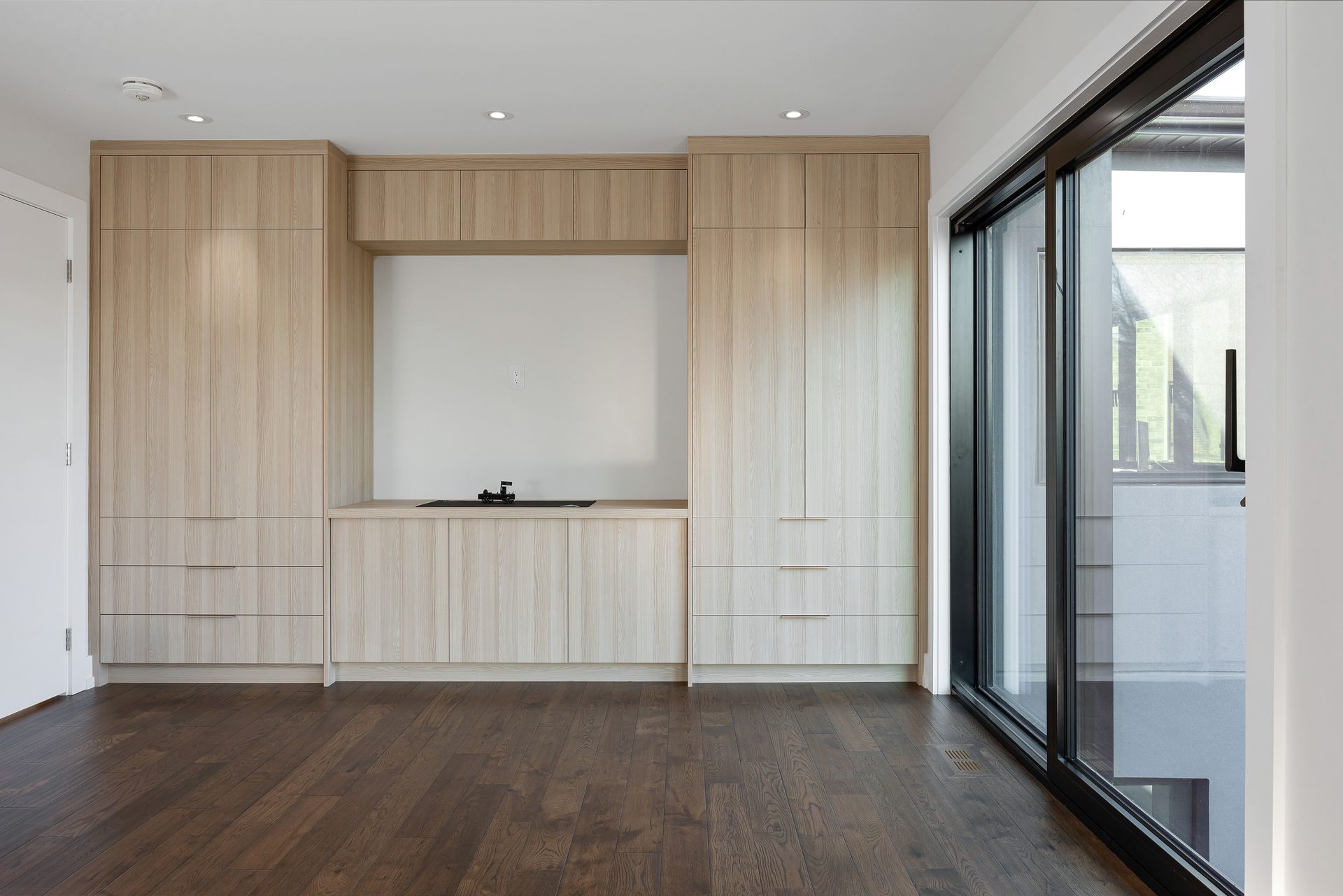 Modern empty room with built-in light wood cabinets, dark floor, and sliding glass doors on the right