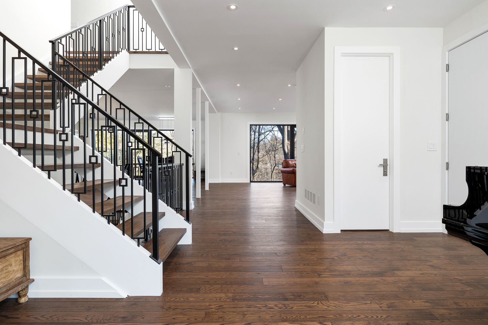 Bright foyer with hardwood floors, staircase, white walls, and a long hallway opening to a distant doorway.