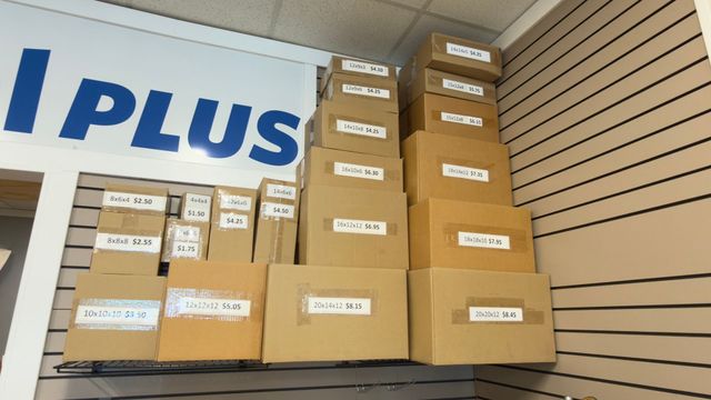 Stacked cardboard shipping boxes in a delivery truck, with an I-PLUS sign on the wall.