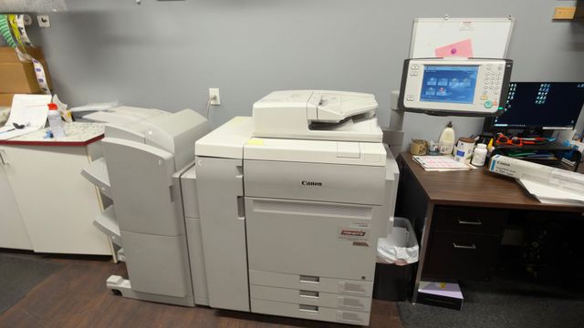 Office copy room with large printer, desk, computer monitor, and scattered papers.