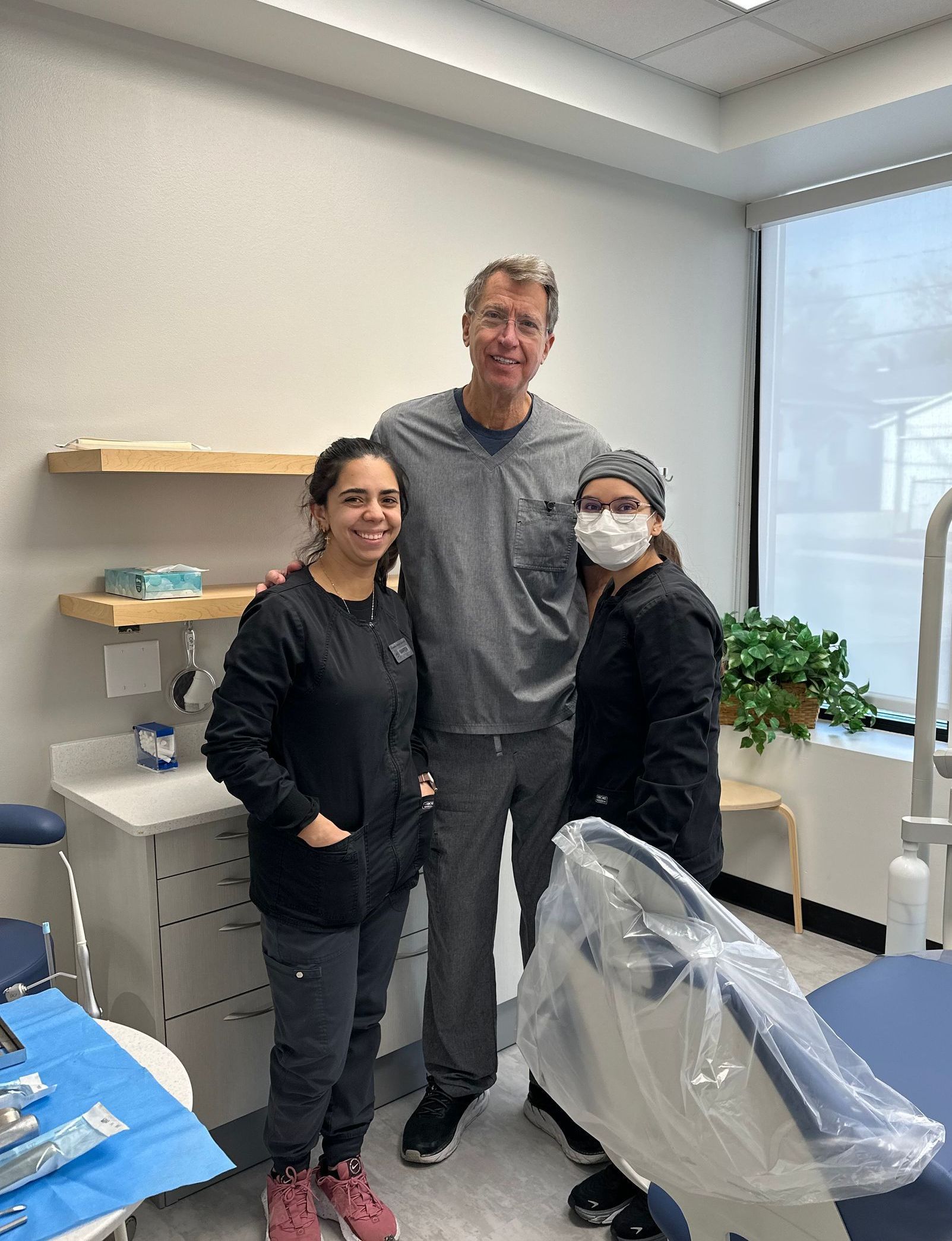 Three people standing together in a clinic room, smiling beside exam equipment and a window.
