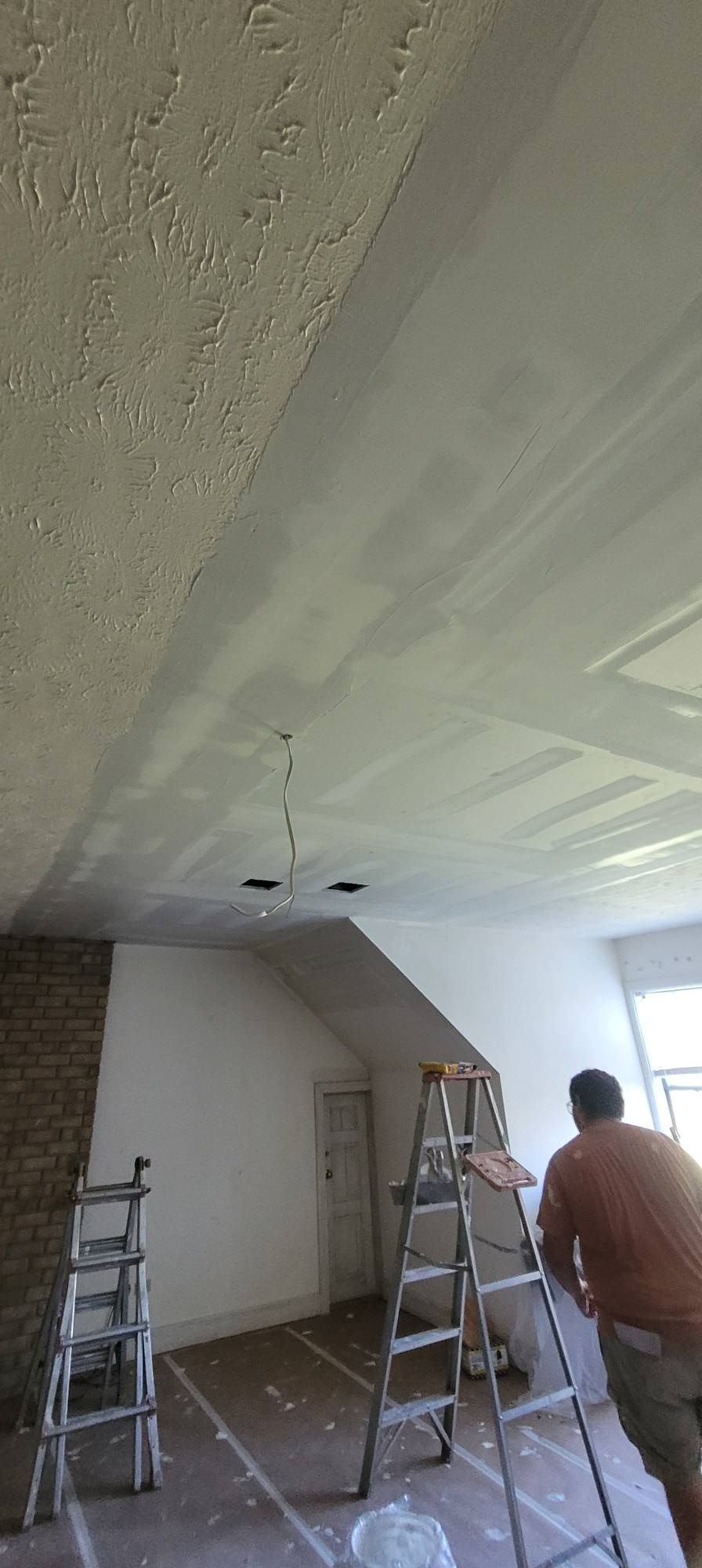 A person working on a ceiling with two ladders in a room under renovation. One section of the ceiling has popcorn texture.