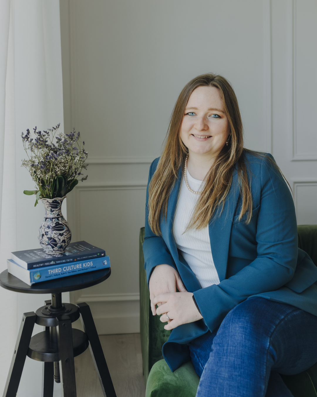 Woman seated in a teal chair beside a side table with books and a vase of flowers in a bright room