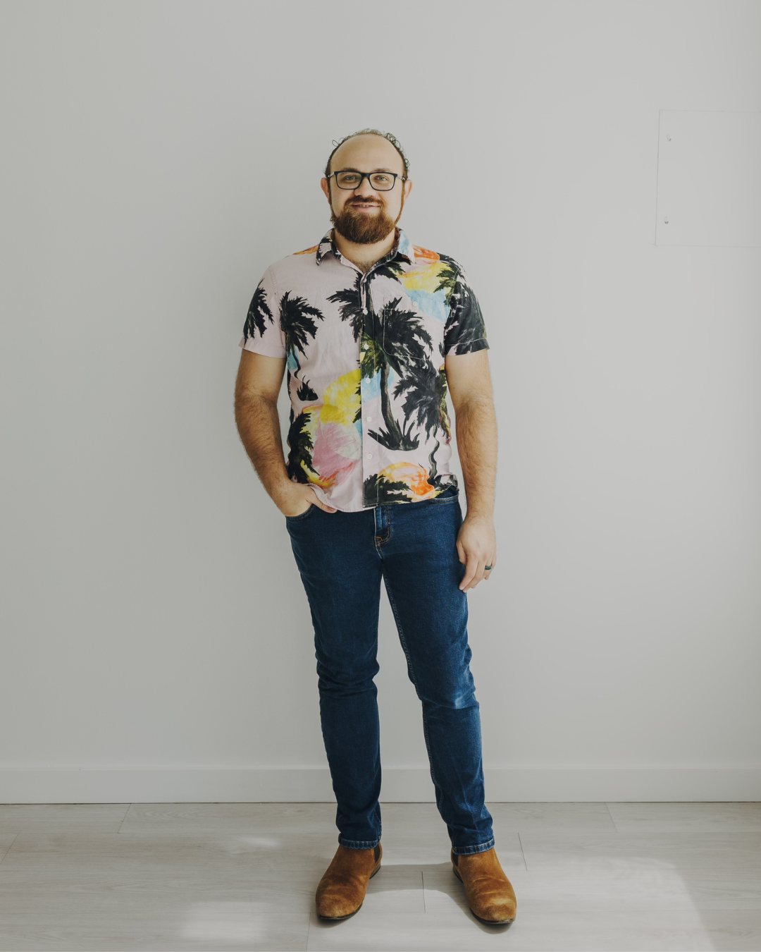 Man standing against a plain wall, wearing a floral shirt, jeans, and brown shoes.