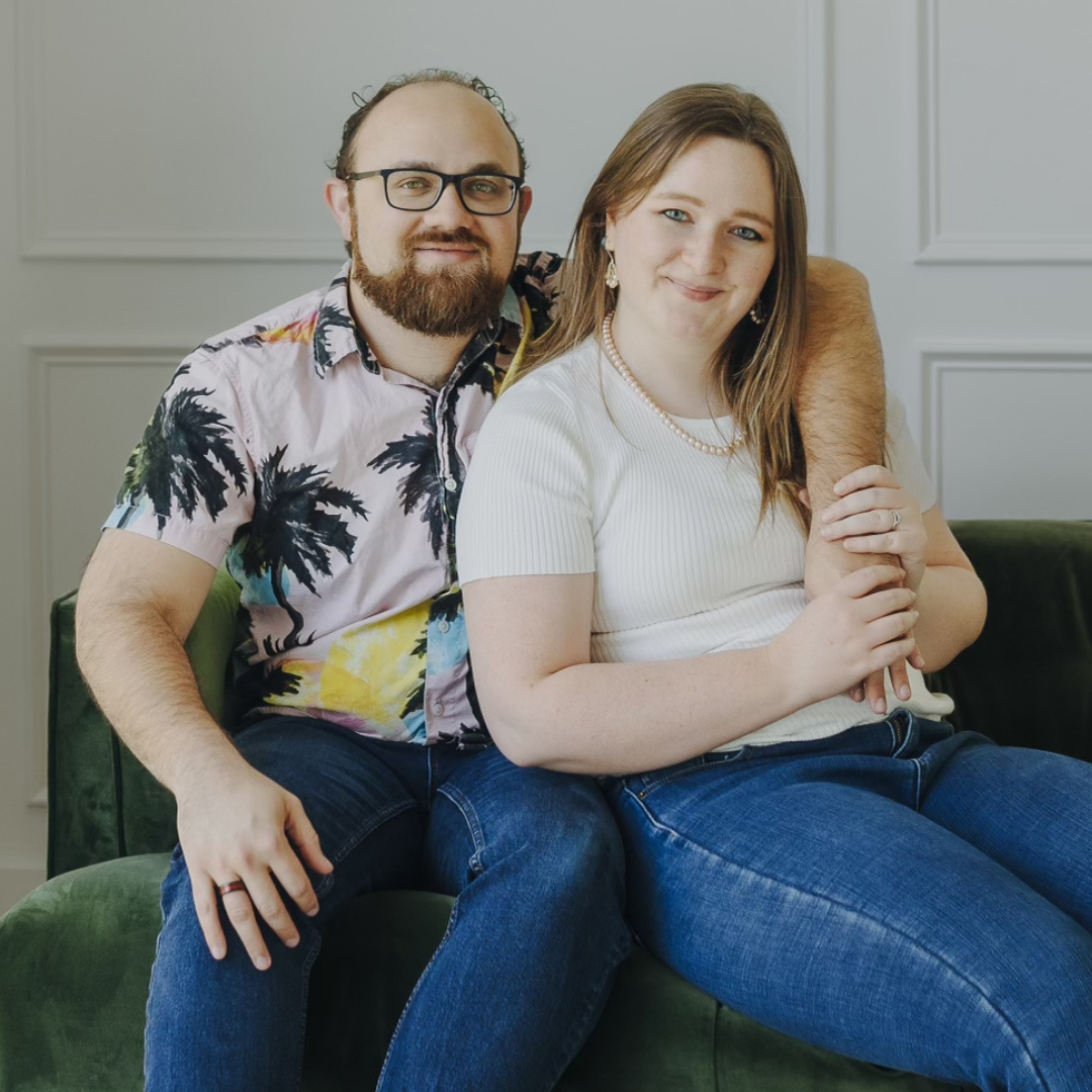 Two people sitting together on a couch, smiling at the camera in a bright living room.