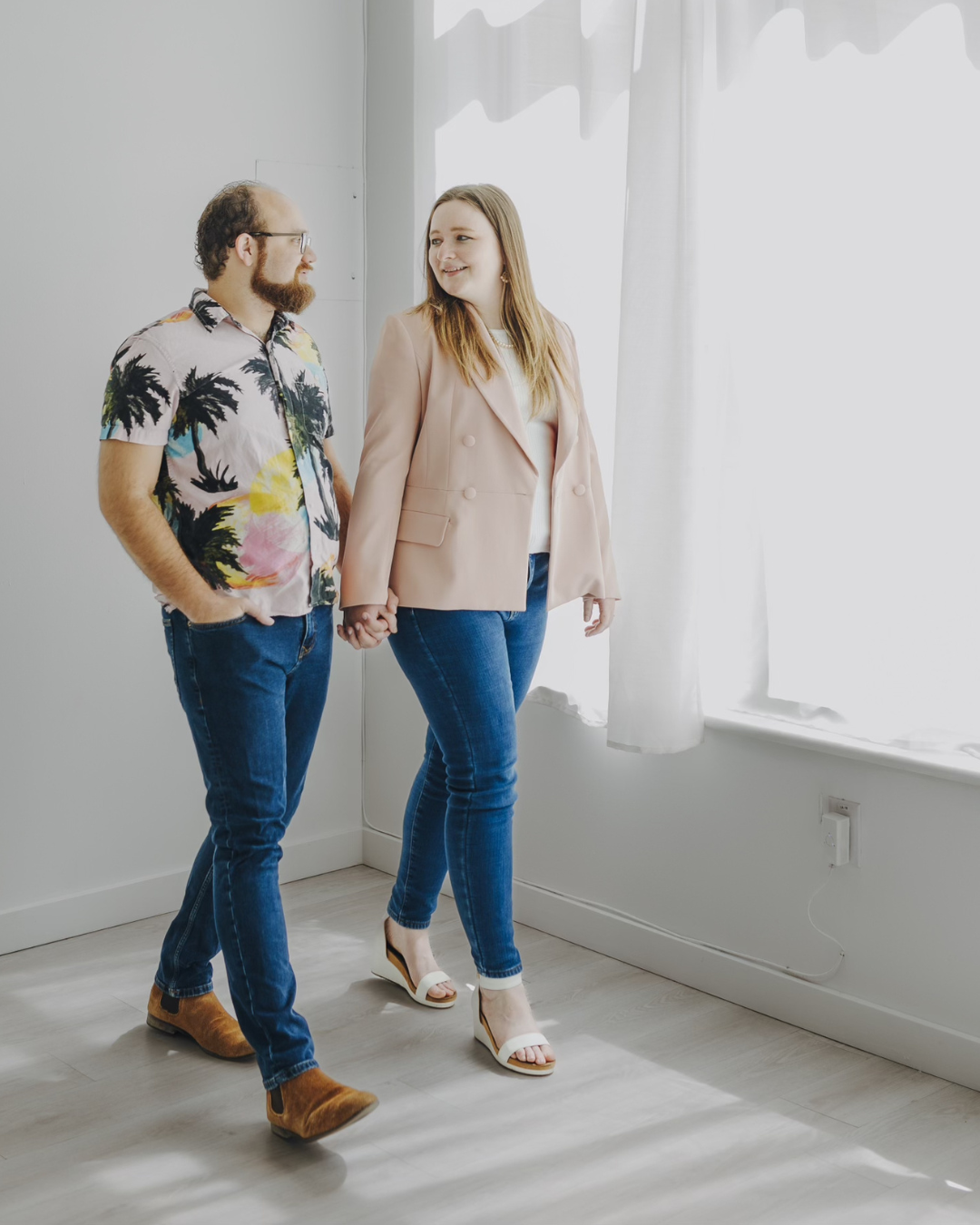 Two people walking hand in hand in a bright, empty room near large windows.