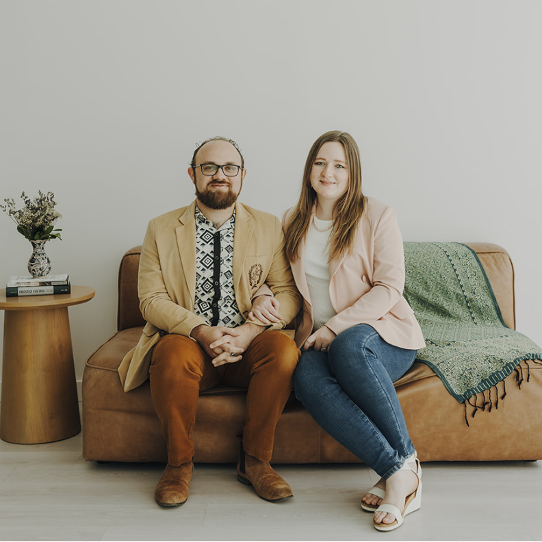 Two people sit on a tan couch in a bright room, with a small table and flowers beside them.