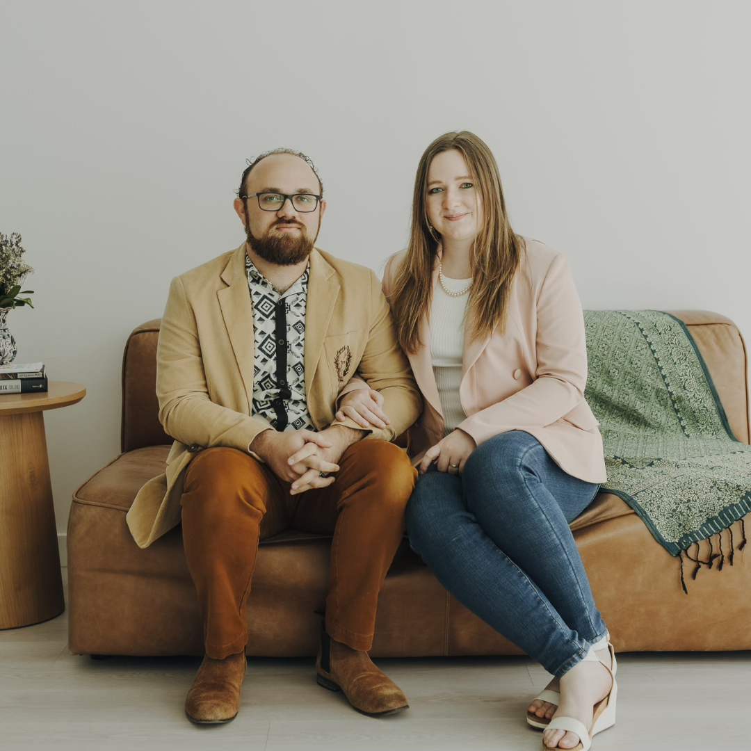 Two people sitting on a couch in a bright room, one in a tan jacket and one in a pink blazer