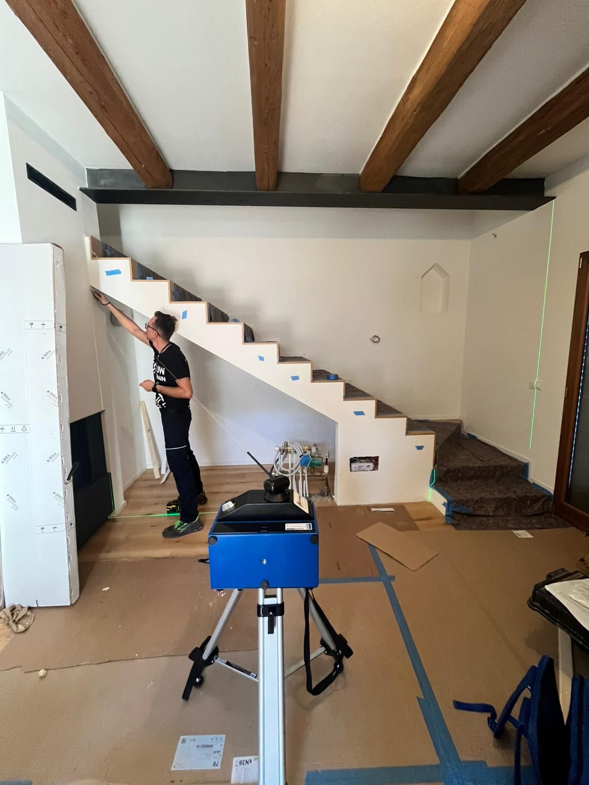 Una persona lavora su una scala di legno in una stanza con travi a vista sul soffitto e carta protettiva sul pavimento; un treppiede si trova nelle vicinanze.