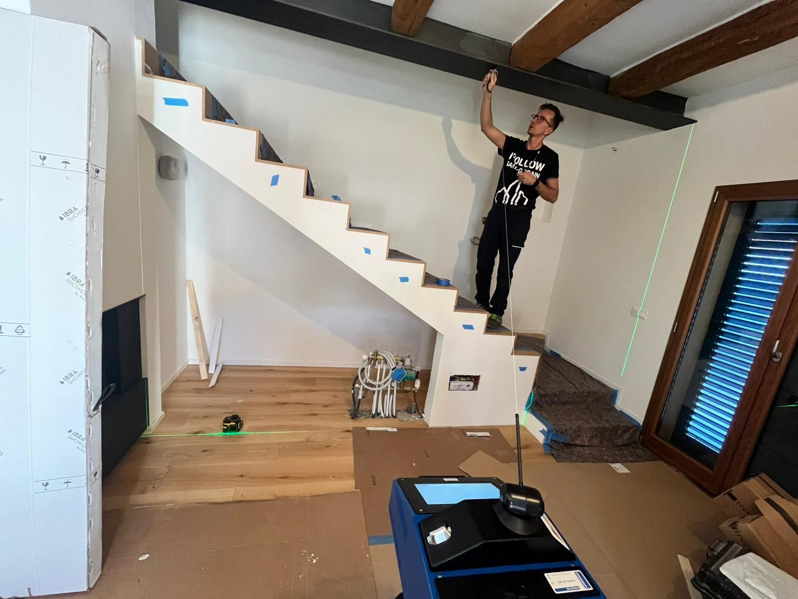 Una persona si trova su una scala di legno in una stanza in costruzione e utilizza una livella laser per misurare una trave nera sul soffitto.
