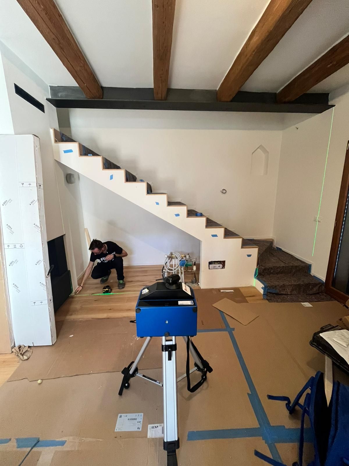 Una persona lavora su un pavimento di legno vicino a una scala, con uno scanner laser 3D su un treppiede in primo piano.