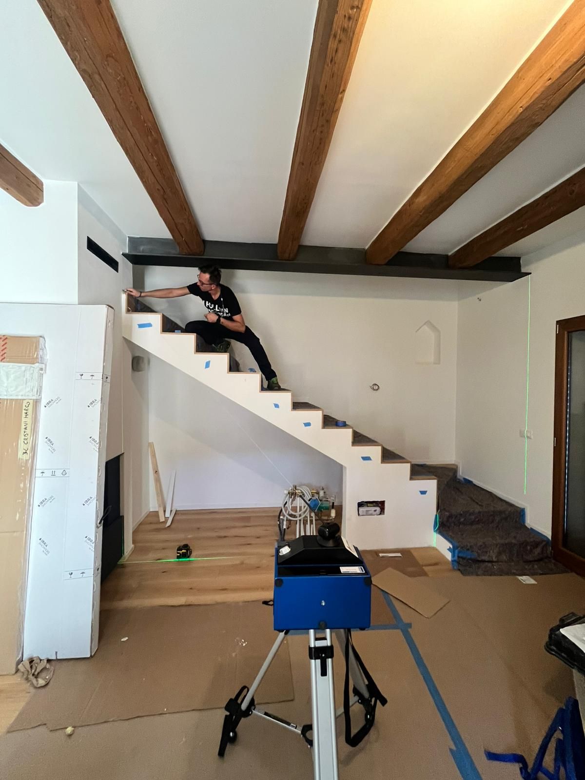 Una persona installa un corrimano per scale in una stanza con travi a vista sul soffitto e rivestimenti protettivi sul pavimento.