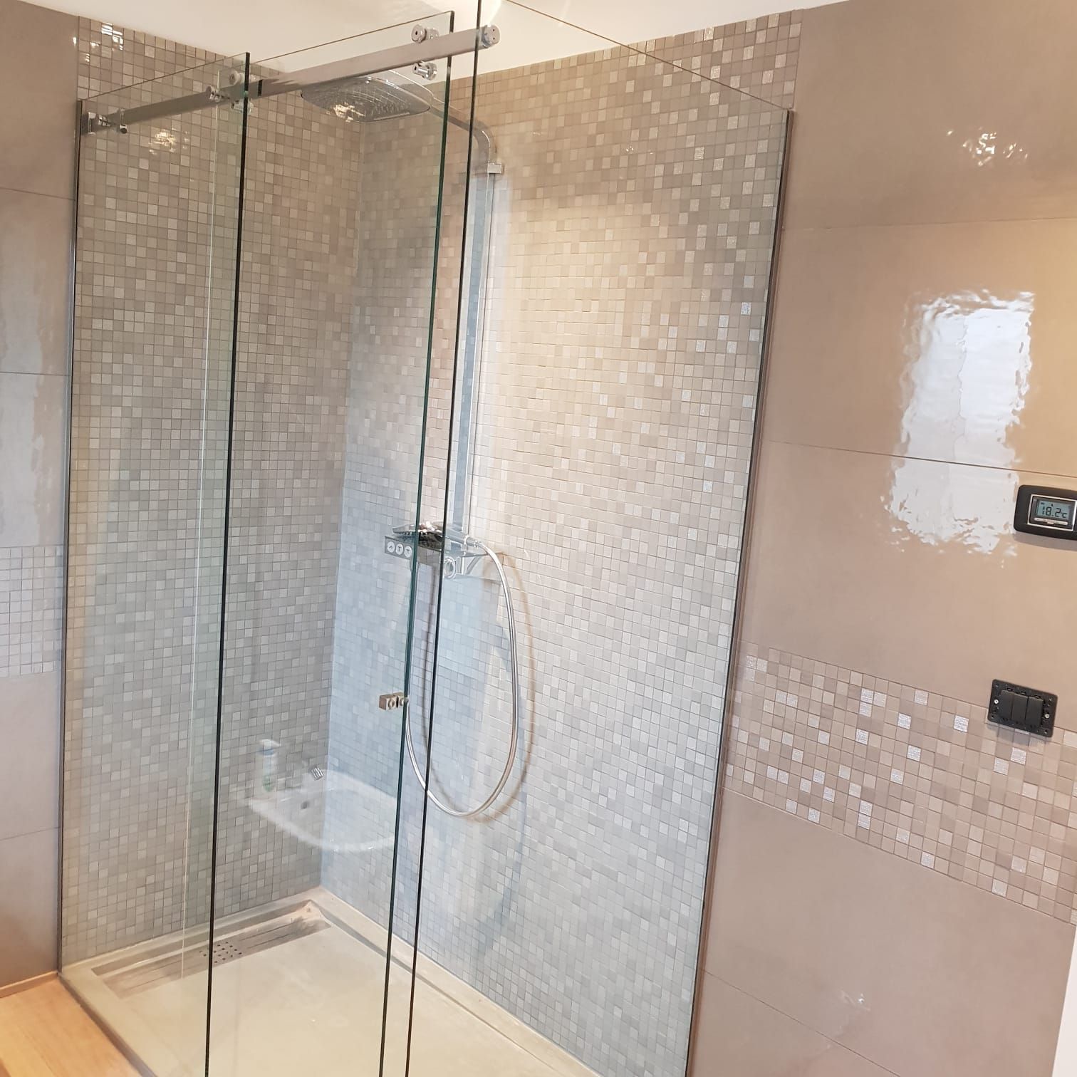 Una moderna cabina doccia con box in vetro, pareti rivestite con piastrelle a mosaico di colore chiaro e piatto doccia ribassato.