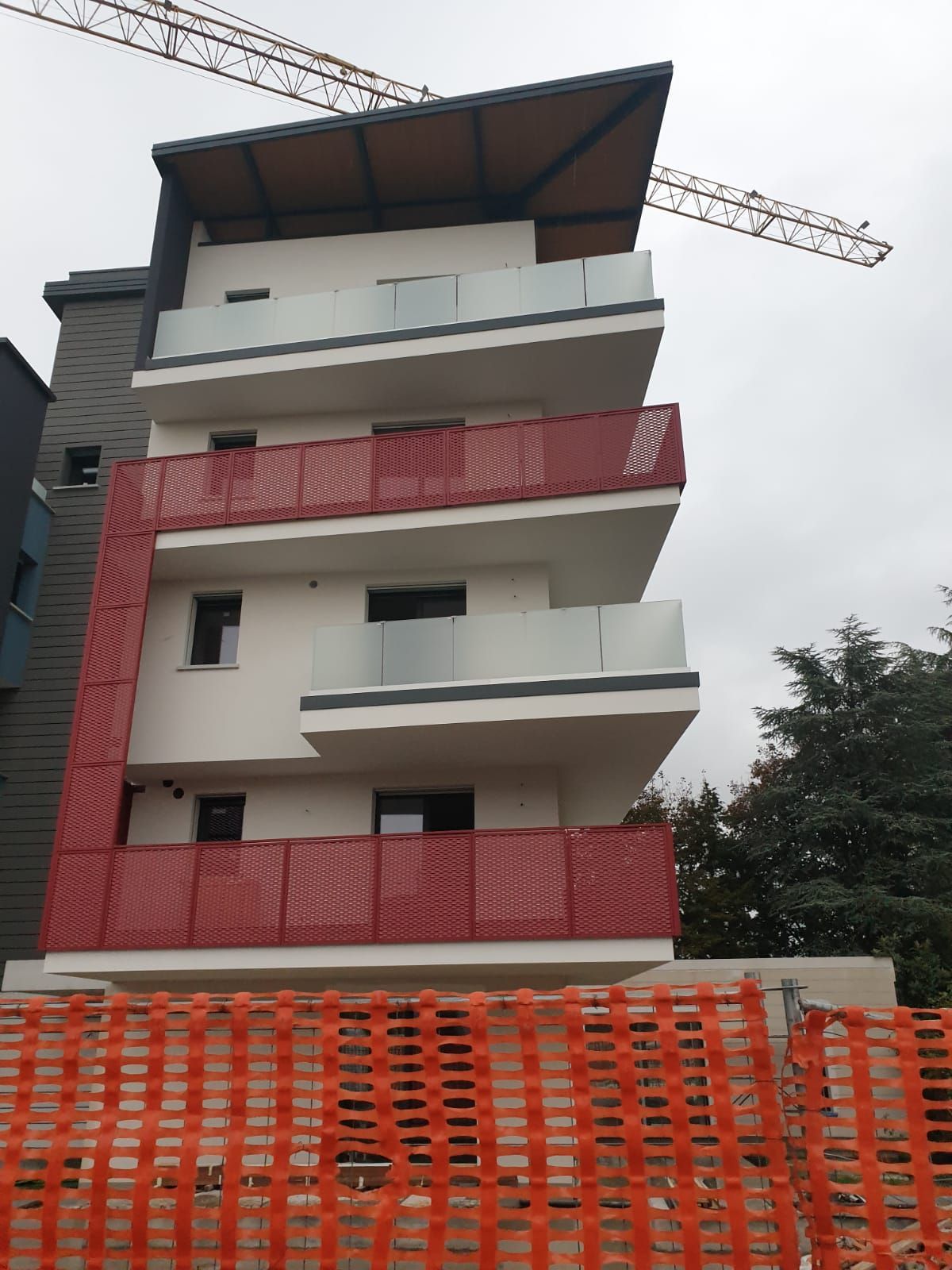 Moderno condominio con balconi bianchi sfalsati, ringhiere in rete metallica rossa e una gru che si staglia nel cielo nuvoloso.