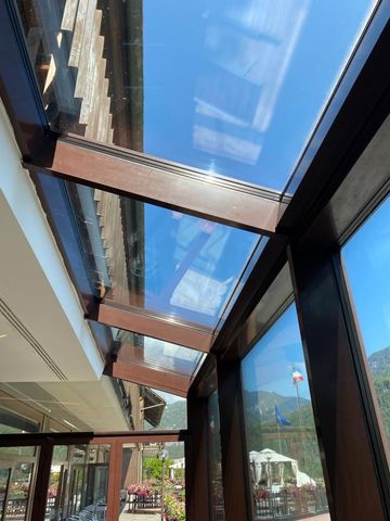 Una vista attraverso una serra con tetto in vetro e struttura in legno marrone scuro, che si affaccia su un paesaggio soleggiato e sulle colline.