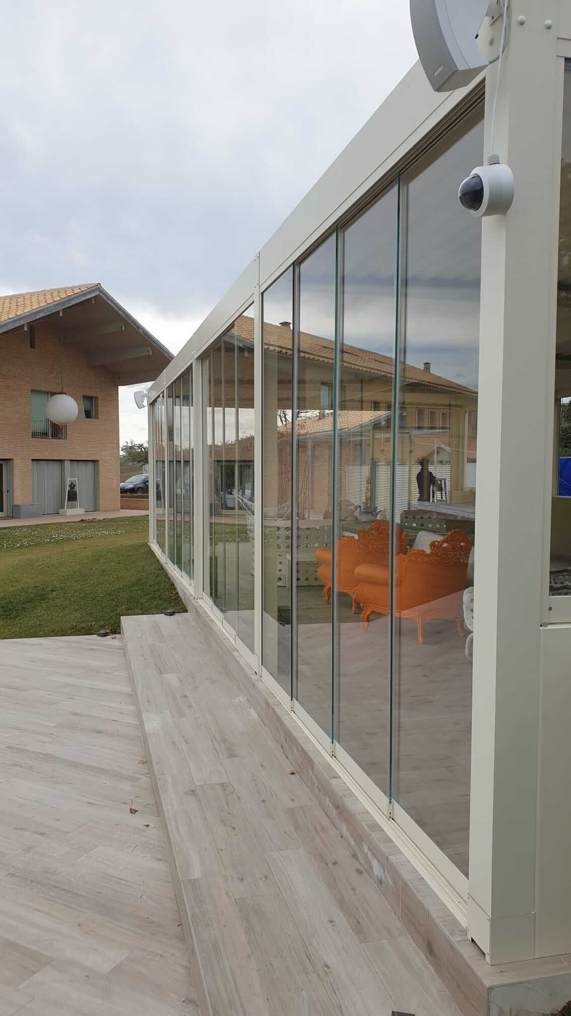 Una moderna struttura esterna con pareti in vetro e telaio bianco, situata su un patio adiacente a una casa con facciata in legno.