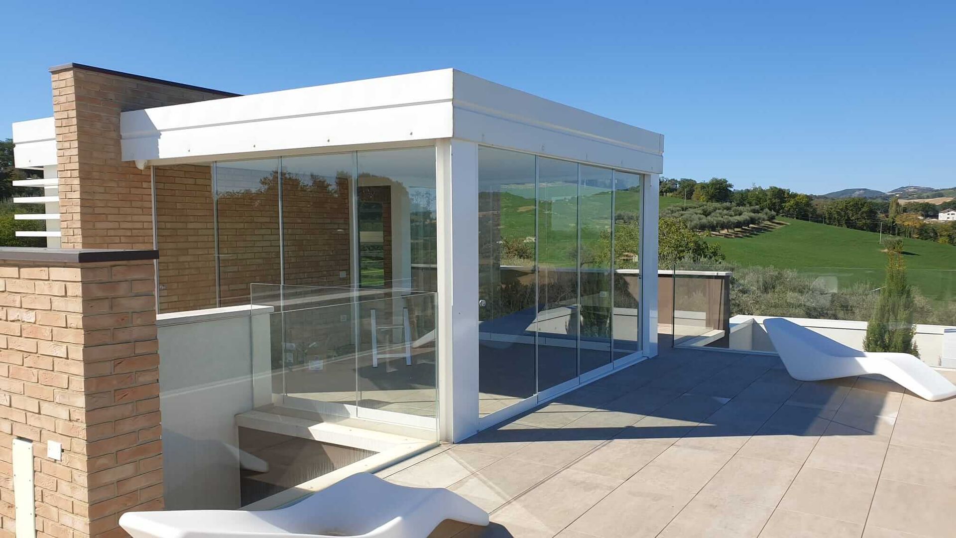 Una moderna struttura in vetro con cornice bianca su un patio piastrellato, con un muro in mattoni e sedie a sdraio da esterno sotto un cielo limpido.