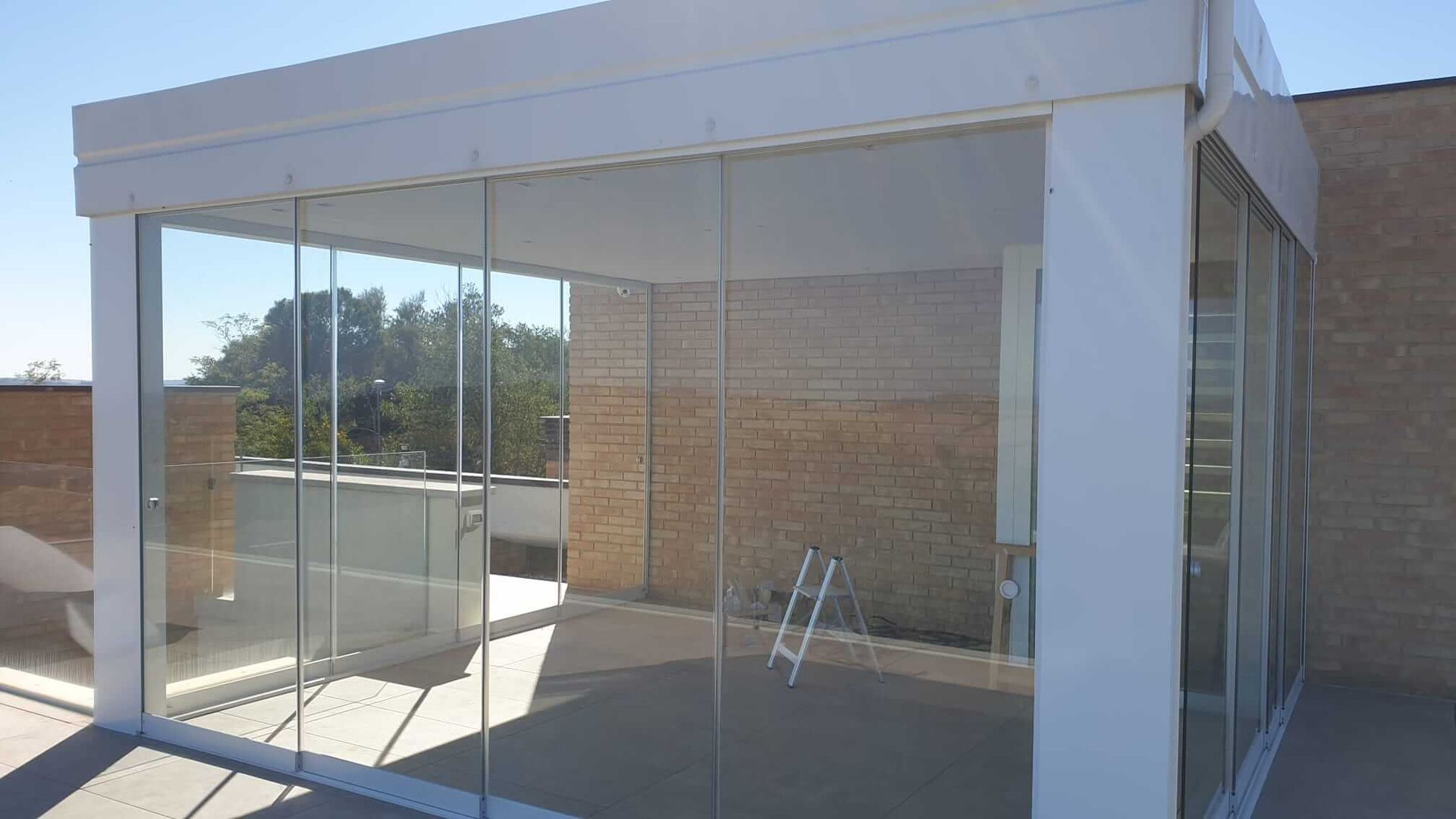 In una giornata di sole, su una terrazza sul tetto, addossata a un edificio in mattoni, si erge una moderna struttura da esterno con pareti in vetro.