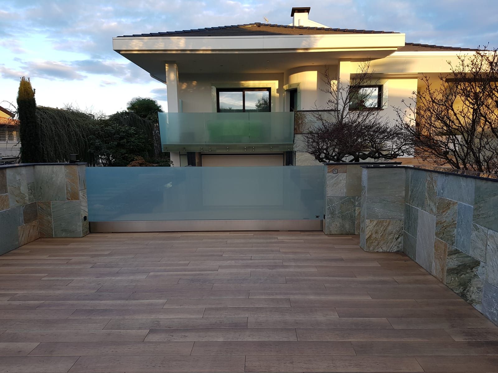 Una terrazza in legno conduce a una casa moderna con muri in pietra e un cancello in vetro satinato, vista in una giornata di sole.