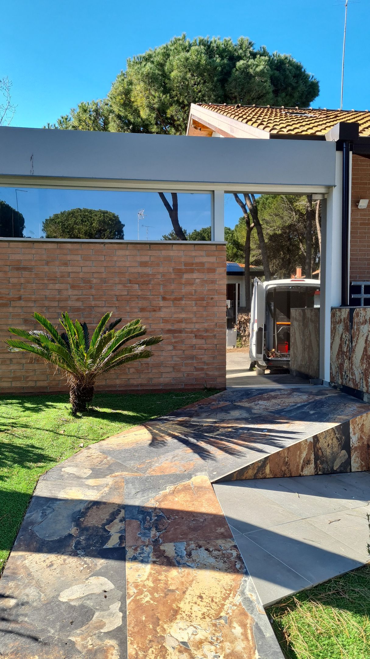 Un sentiero di pietra conduce all'ingresso di un cortile con un muro di mattoni, una piccola palma e una vista parziale di un veicolo parcheggiato.