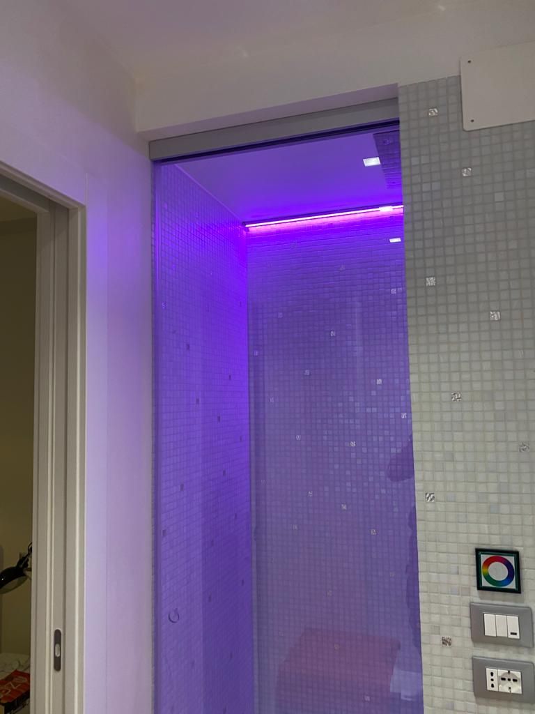 Un box doccia con un'illuminazione d'accento a LED viola brillante installata vicino al soffitto, fiancheggiato da pareti rivestite di piastrelle bianche.