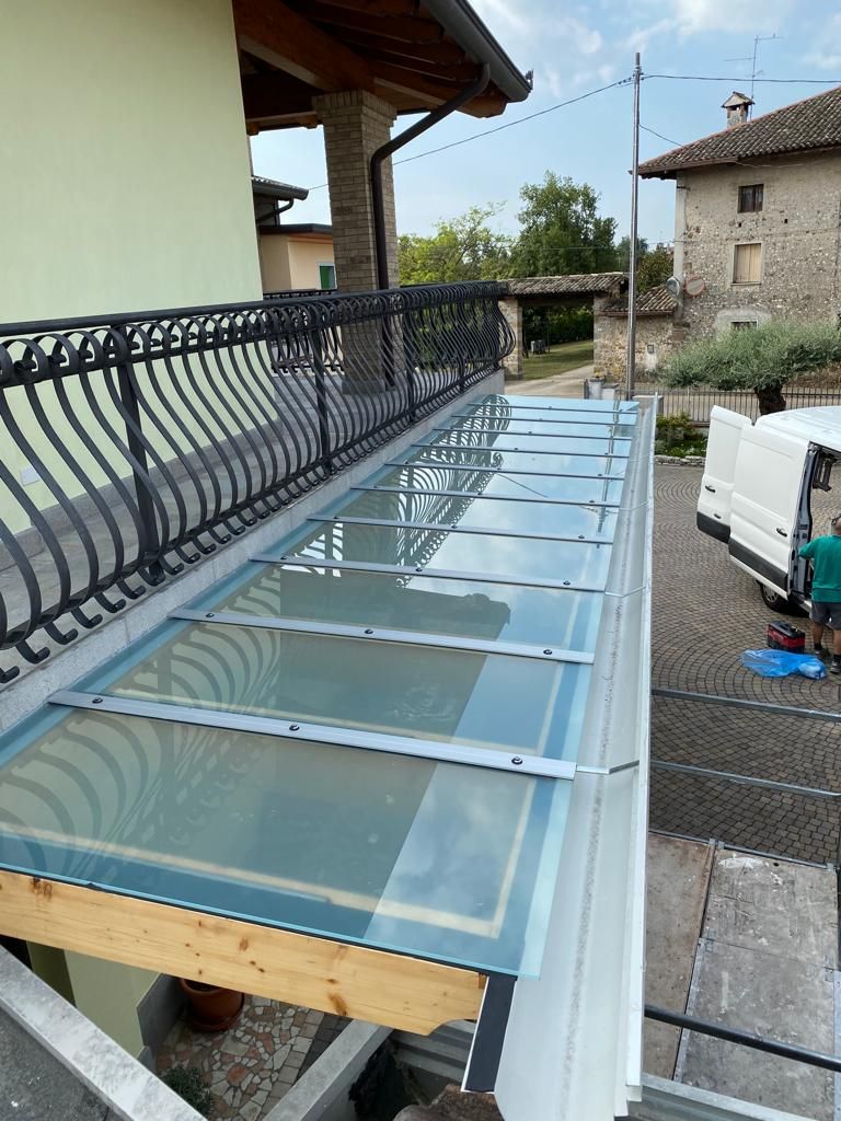 Una tettoia in vetro di recente installazione per un balcone, con travi di supporto in metallo, vista dall'alto accanto a un edificio.