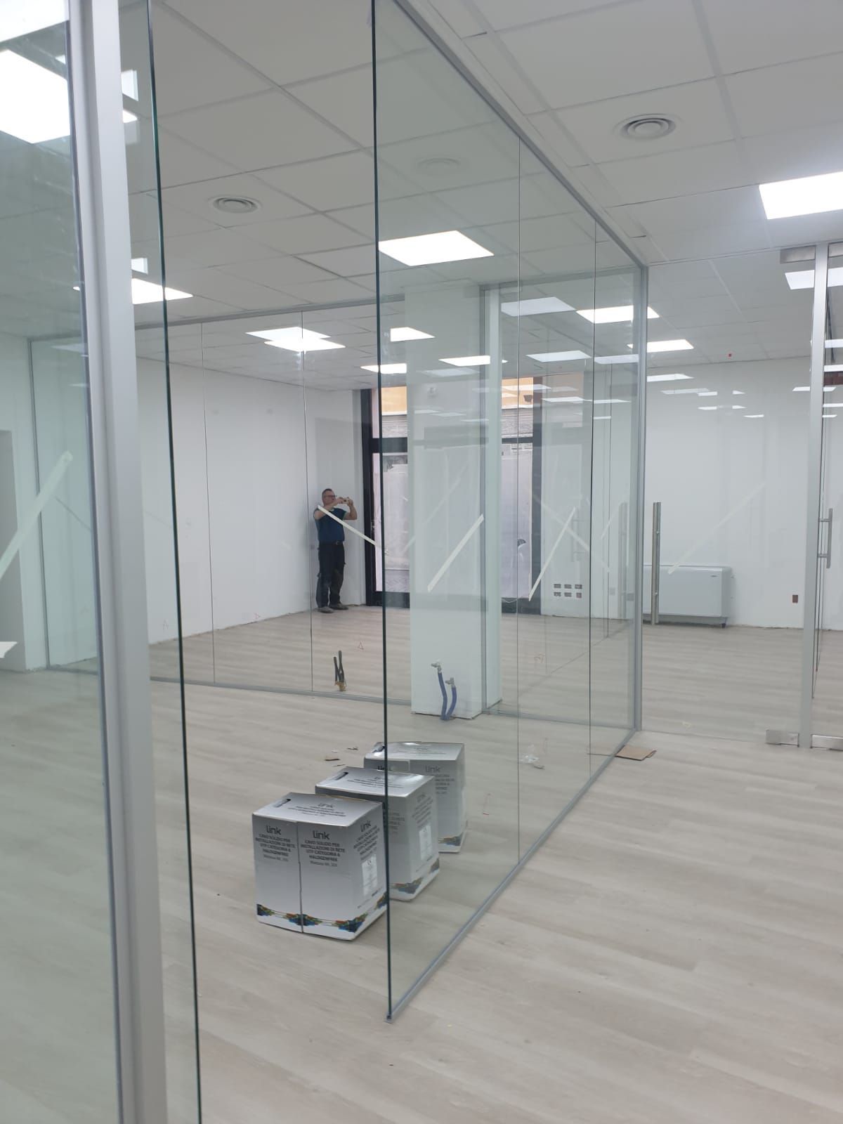 Vista interna di un ufficio con pareti divisorie in vetro e pavimento moderno e chiaro, illuminato da luminose luci a soffitto.