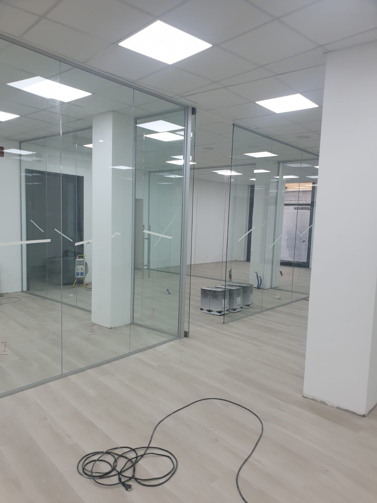 Un ufficio open space caratterizzato da pareti bianche, divisori in vetro, luci a soffitto e un cavo nero appoggiato sul pavimento.