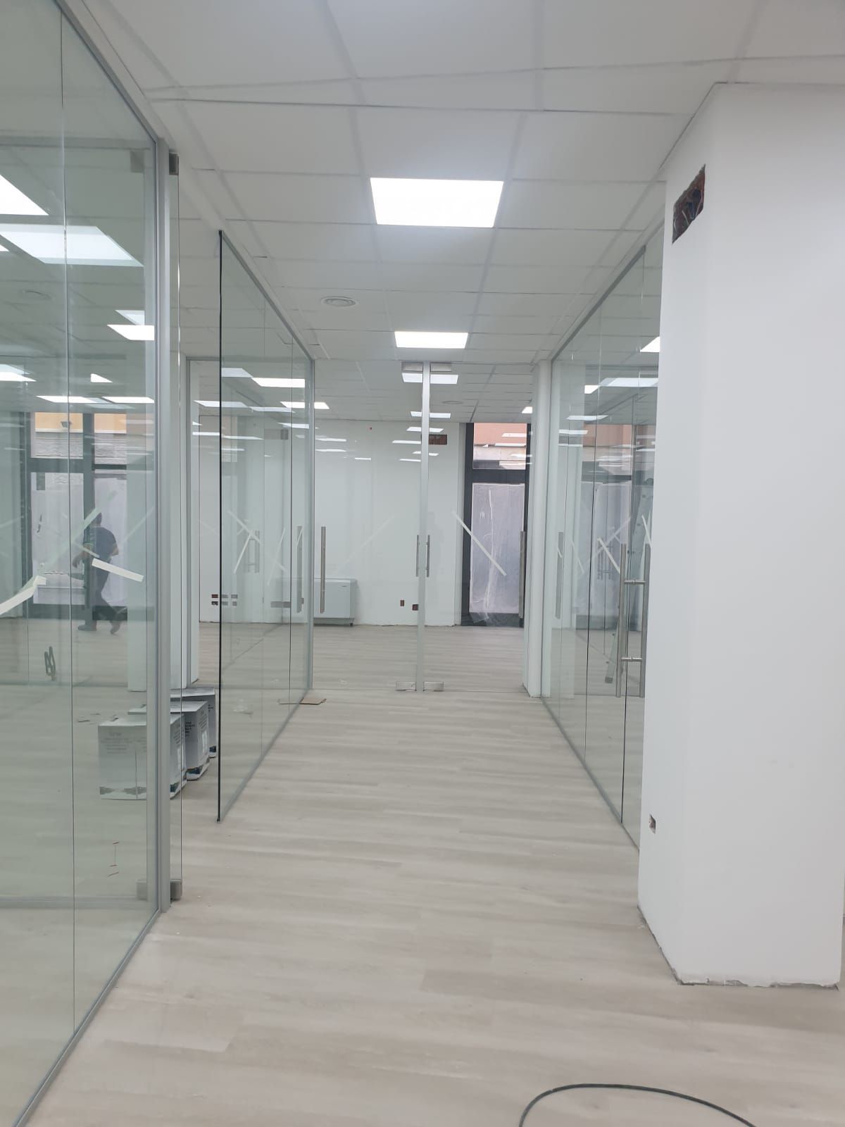 Un corridoio per uffici luminoso e moderno, con pareti divisorie in vetro su entrambi i lati, pavimenti in legno chiaro e una colonna bianca sulla destra.