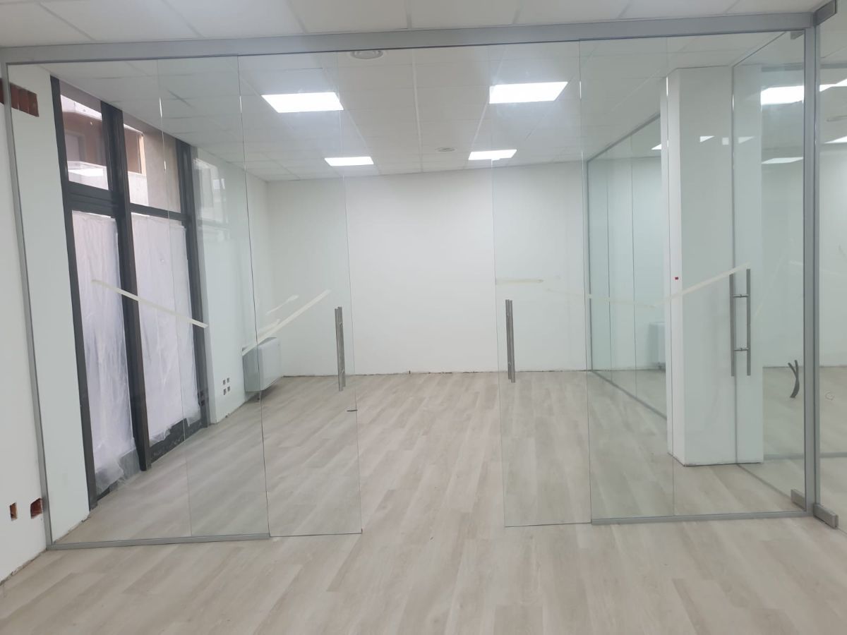 Un ufficio vuoto e luminoso, con pavimento in legno chiaro e ampie pareti divisorie in vetro.