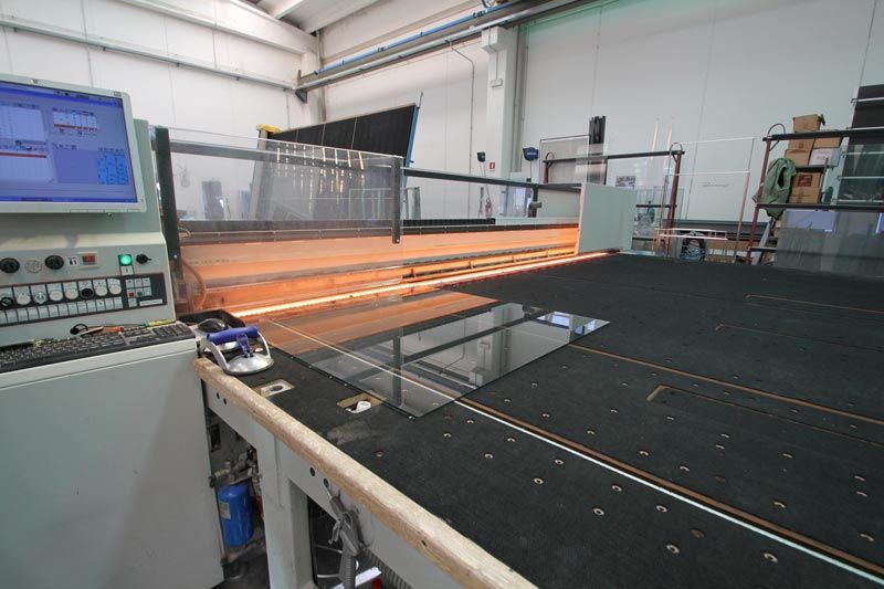 Un tavolo da taglio per vetro a controllo numerico (CNC) in una fabbrica, dotato di una superficie di lavoro piana e di un braccio robotico che scansiona una lastra di vetro scuro.