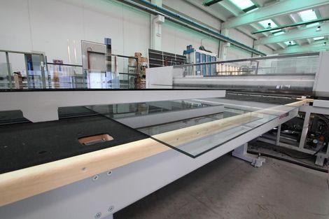 Una lastra di vetro piatta appoggiata su un tavolo da taglio industriale all'interno di uno stabilimento di produzione ben illuminato.