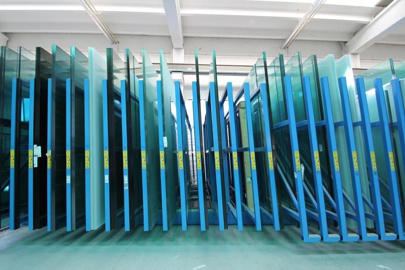 Grandi lastre di vetro industriale sono stoccate in posizione verticale su scaffalature metalliche blu ben organizzate all'interno di un magazzino luminoso.