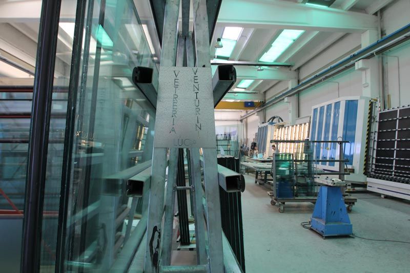 L'officina è caratterizzata da alte scaffalature metalliche a forma di A che sorreggono pannelli di vetro, con macchinari industriali e postazioni di lavoro nella parte posteriore.