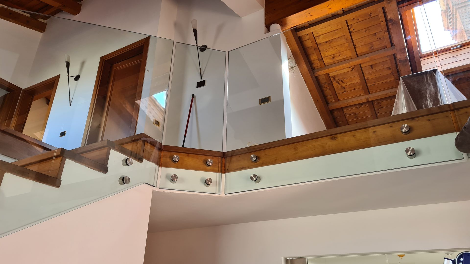Un balcone in legno con ringhiera in vetro fissata tramite supporti metallici, che si affaccia su uno spazio interno con soffitto inclinato.