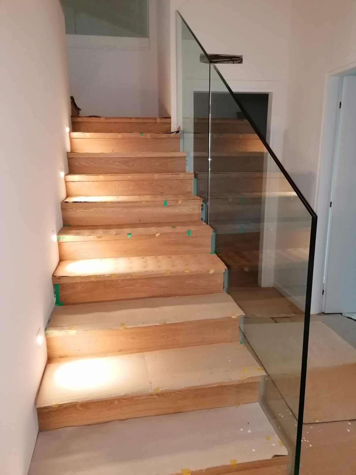 Scala in legno chiaro che sale verso l'alto, con ringhiera moderna in vetro senza telaio e applique a parete sulla sinistra.