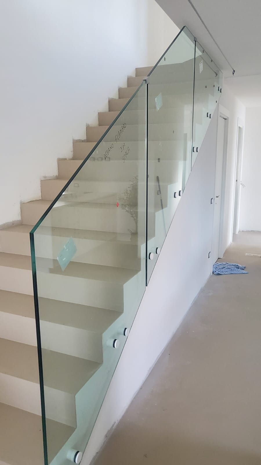 Una scala con gradini beige e una ringhiera in vetro senza telaio, fissata lateralmente con elementi metallici.