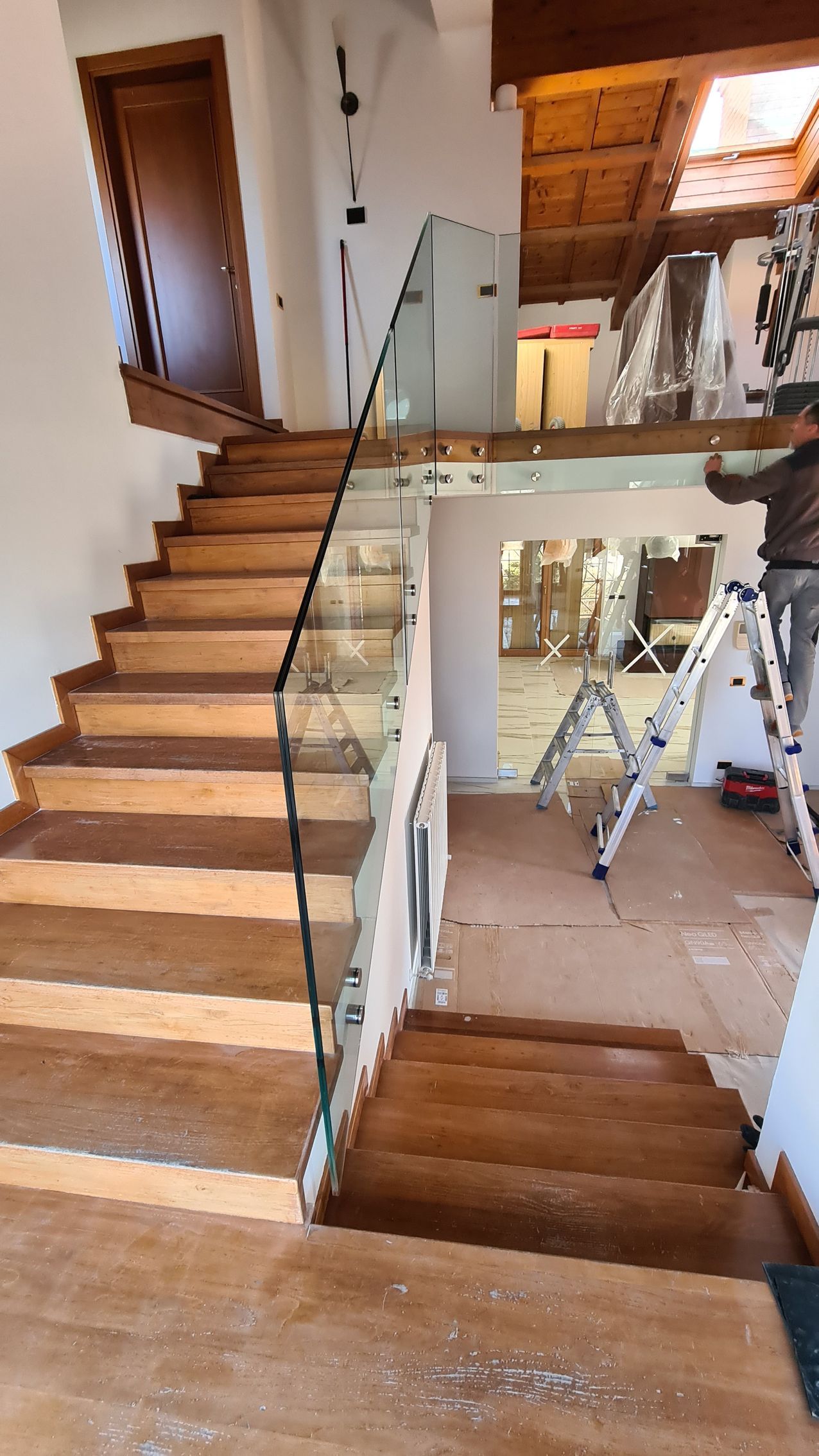 Una scala con gradini in legno e ringhiera in vetro, che conduce a un piano superiore in costruzione.